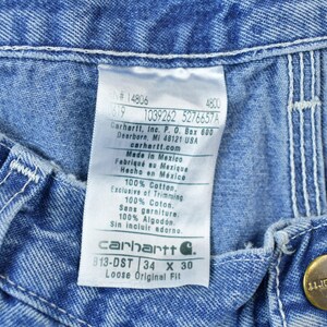 May include: A blue denim Carhartt label with white text. The label reads "#14806 4800 619 1039262 5276657A Carhartt, Inc. P.O. Box 600 Dearborn, MI 48121 USA carhartt.com Made in Mexico Fabriqu&eacute; au Mexique Hecho en M&eacute;xico 100% Cotton. Exclusive of Trimming 100% Cotos. Sans garniture. 100% Algod&oacute;n Sin incluir adorno. carhartt G. 813-DST 34 X 30 Loose Original Fit"