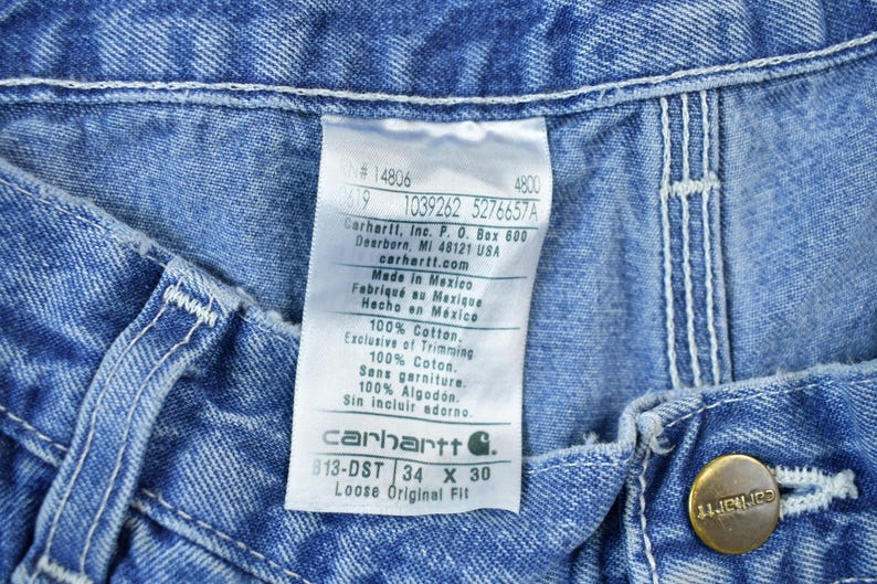 May include: A blue denim Carhartt label with white text. The label reads "#14806 4800 619 1039262 5276657A Carhartt, Inc. P.O. Box 600 Dearborn, MI 48121 USA carhartt.com Made in Mexico Fabriqu&eacute; au Mexique Hecho en M&eacute;xico 100% Cotton. Exclusive of Trimming 100% Cotos. Sans garniture. 100% Algod&oacute;n Sin incluir adorno. carhartt G. 813-DST 34 X 30 Loose Original Fit"