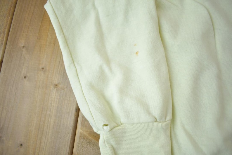 May include: A pale yellow sweatshirt with a small brown stain on the front. The sweatshirt has long sleeves and a ribbed cuff.