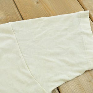 May include: A short-sleeved t-shirt in a solid off-white or cream color. The garment features a standard crew neck construction with visible stitching along the sleeve hem and shoulder seam, showcasing a plain, unpatterned fabric texture.