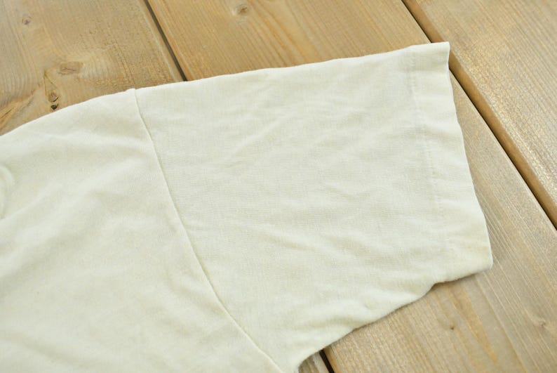 May include: A short-sleeved t-shirt in a solid off-white or cream color. The garment features a standard crew neck construction with visible stitching along the sleeve hem and shoulder seam, showcasing a plain, unpatterned fabric texture.