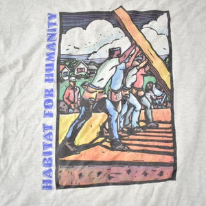 May include: A vintage grey t-shirt featuring a colourful illustration of people building a house. The text "HABITAT FOR HUMANITY" is printed on the left side of the shirt.