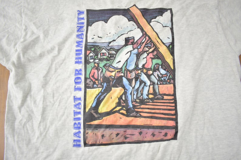 May include: A vintage grey t-shirt featuring a colourful illustration of people building a house. The text "HABITAT FOR HUMANITY" is printed on the left side of the shirt.
