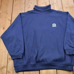 Könnte beinhalten: Ein marineblauer Rollkragenpullover mit einem weißen Logo auf der Vorderseite. Das Logo zeigt eine Krone und den Text "The Crown Collection".