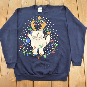Puede incluir: Una sudadera azul marino de manga larga con un gráfico de un gato blanco con astas de reno. El gato está rodeado por una cadena de luces navideñas de colores y puntos blancos dispuestos en forma de árbol de Navidad.