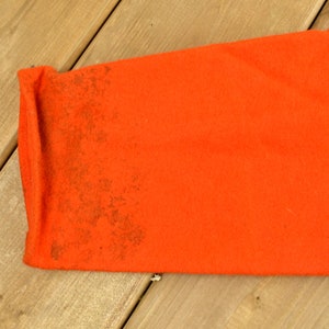 K&ouml;nnte beinhalten: Nahaufnahme des orangenen Stoff&auml;rmels eines Kleidungsst&uuml;cks. Der Stoff hat eine strukturierte, leicht abgenutzte Optik mit braunen Flecken.
