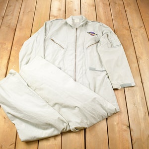May include: A light gray flight suit with a zipper closure and a patch on the left chest. The suit is folded and laid out on a wooden surface.