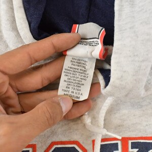 Op de afbeelding: Een lichtgrijze gem&ecirc;leerde hoodie met een donkerblauwe voering in de capuchon en een wit trekkoord. Het kledingstuk heeft een wit waslabel met zwarte tekst die 50% katoen en 50% Fortrel-polyester aangeeft, in maat medium.