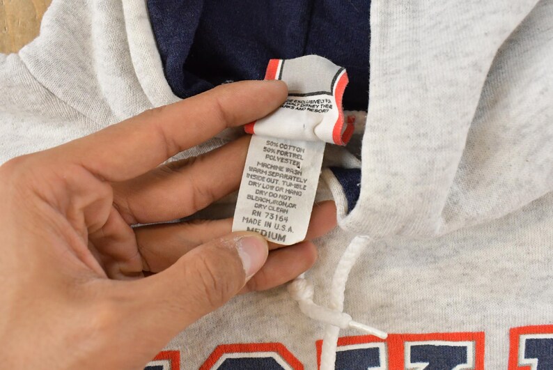 Op de afbeelding: Een lichtgrijze gem&ecirc;leerde hoodie met een donkerblauwe voering in de capuchon en een wit trekkoord. Het kledingstuk heeft een wit waslabel met zwarte tekst die 50% katoen en 50% Fortrel-polyester aangeeft, in maat medium.