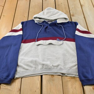 May include: A long-sleeve pullover hoodie featuring a colour-blocked design in blue, light grey, and maroon. The garment includes a hood with drawstrings, a partial button-up placket at the neckline, and a horizontal flap detail across the chest.