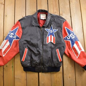 Puede incluir: Una chaqueta bomber de cuero negro con mangas rojas y azules. Las mangas presentan una gran estrella con la palabra "USA" en su interior. La chaqueta tiene una cremallera y un cuello de canalé.