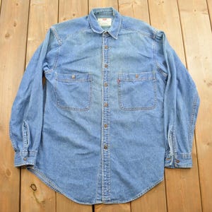 Pode incluir: Uma camisa de brim azul claro de manga comprida com colarinho de botões e dois bolsos no peito. A camisa tem um logotipo vermelho Levi's no bolso.