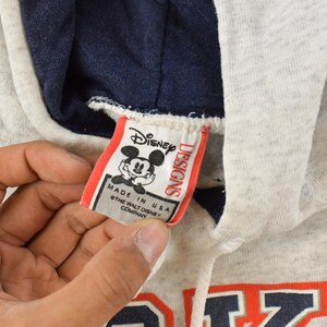 Op de afbeelding: Een lichtgrijze gem&ecirc;leerde hoodie met een donker marineblauwe voering in de capuchon en een wit trekkoord. Een wit stoffen label toont het Disney Designs logo met een Mickey Mouse afbeelding en de tekst Made in USA en copyright.