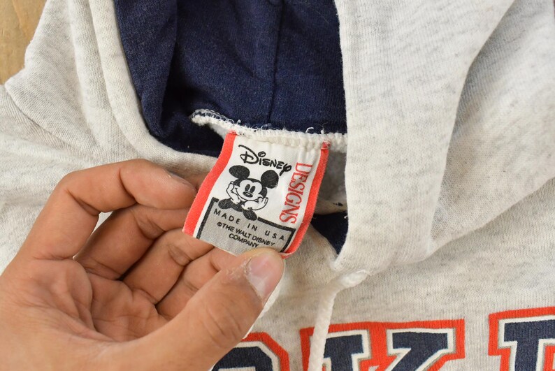 Op de afbeelding: Een lichtgrijze gem&ecirc;leerde hoodie met een donker marineblauwe voering in de capuchon en een wit trekkoord. Een wit stoffen label toont het Disney Designs logo met een Mickey Mouse afbeelding en de tekst Made in USA en copyright.