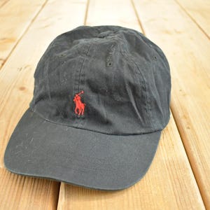 Könnte beinhalten: Eine schwarze Baseballkappe mit gebogenem Schirm. Auf der Kappe befindet sich ein rotes, gesticktes Logo eines Polospielers. Der Hut ist aus einem weichen, abgenutzten Stoff gefertigt.