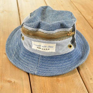 Könnte beinhalten: Ein blauer Jeans-Anglerhut mit einem horizontalen Messing-Reißverschluss um die Krone, zwei Belüftungsösen aus Messing und einem weißen, rechteckigen Stoffaufnäher mit dem Text Casuals with Class Made in U.S.A. auf der Vorderseite.