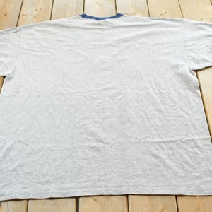 Puede incluir: Una camiseta gris jaspeado con cuello y ribetes de manga azul marino. La camiseta est&aacute; tendida sobre una superficie de madera. La camiseta es de estilo cuello redondo.