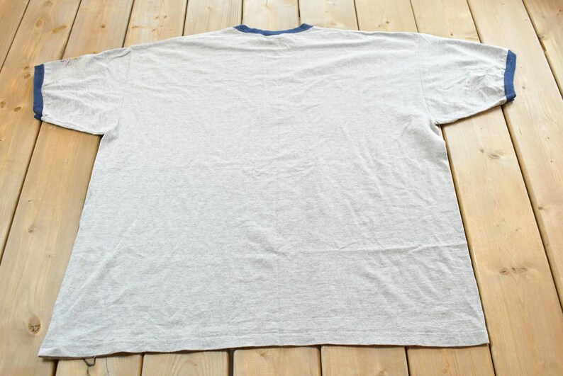 Puede incluir: Una camiseta gris jaspeado con cuello y ribetes de manga azul marino. La camiseta est&aacute; tendida sobre una superficie de madera. La camiseta es de estilo cuello redondo.