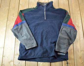 Puede incluir: Un jersey de forro polar azul con cierre de cremallera de media cremallera. Las mangas son una mezcla de gris, verde, rojo y azul.