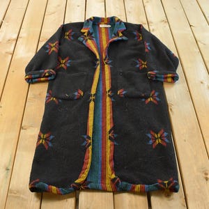 Peut inclure: Un long manteau noir à manches longues avec des motifs floraux et géométriques colorés. Le manteau présente une bande verticale de couleurs arc-en-ciel au centre et sur le col. Les poignets et les poches sont également ornés de motifs colorés.