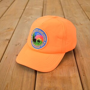 May include: Vibrant orange baseball cap with a curved peak. The cap has a circular patch depicting a mountain scene, deer silhouettes, and the text "Colorado Big Game Hunter Anniversary 1999". The cap is made of a sturdy material.