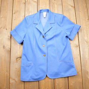 Könnte beinhalten: Ein hellblauer Blazer mit kurzen Ärmeln, Knopfleiste und zwei Taschen.