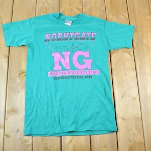 以下が含まれることがあります： ピンク色のグラフィックが特徴のターコイズ色のTシャツ。「NORHTGATE」が「NG」の上に、「HEALTH & RACQUET CLUBS ROCHESTER.MN.」が下に書かれています。半袖のシャツは柔らかい素材で作られています。デザインは幾何学的な形と都市のスカイラインが特徴です。
