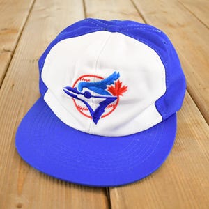 Può includere: Un cappellino da baseball blu e bianco con il logo dei Toronto Blue Jays. Il cappellino presenta una visiera blu e un pannello frontale bianco con il logo della squadra ricamato in blu, rosso e bianco. Il cappellino è probabilmente realizzato in tessuto resistente.