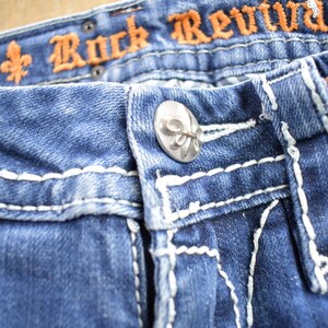K&ouml;nnte beinhalten: Nahaufnahme von blauen Jeans mit wei&szlig;en N&auml;hten und einem silbernen Knopf mit einer Fleur-de-lis-Gravur. Der Bund ist mit den Worten "Rock Revival" in orangefarbenem Garn bestickt.