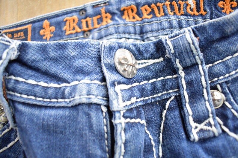 K&ouml;nnte beinhalten: Nahaufnahme von blauen Jeans mit wei&szlig;en N&auml;hten und einem silbernen Knopf mit einer Fleur-de-lis-Gravur. Der Bund ist mit den Worten "Rock Revival" in orangefarbenem Garn bestickt.