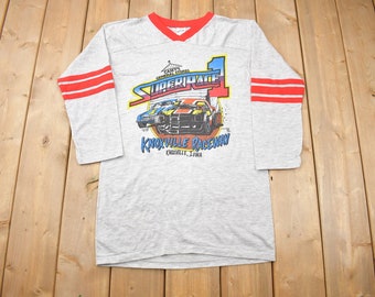 May include: Grey t-shirt with red and white striped sleeves. The shirt features a graphic of two race cars with the text "Super Race 1 Knoxville Raceway Knoxville, Iowa".