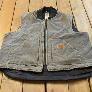 Peut inclure: Un gilet gris Carhartt doté d'un col noir côtelé, d'une fermeture éclair frontale intégrale et de deux grandes poches avant. Le vêtement présente un aspect usé avec une doublure noire matelassée visible à l'ourlet et un petit logo.
