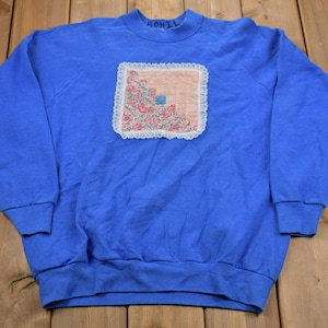 May include: A royal blue sweatshirt with a crew neck and long sleeves. The front features a square patch with a floral design and lace trim. The word "ROHIL" is embroidered on the collar.