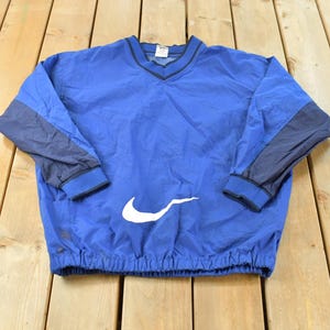 May include: A blue pullover windbreaker jacket featuring a V-neck collar with dark blue trim, long sleeves with dark blue panels, elastic cuffs, and a drawstring hem. A large white Nike swoosh logo is printed on the lower front section.