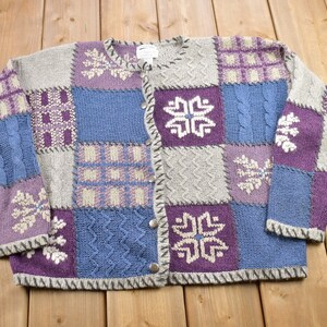 May include: A patchwork cardigan sweater with a button-up front. The sweater is made of a soft, knitted fabric and features a variety of colours, including purple, blue, and grey. The sweater has a snowflake design on the front.