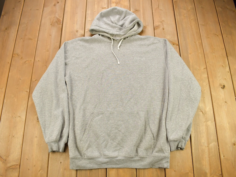 Puede incluir: Una sudadera con capucha gris con un bolsillo canguro en la parte delantera. La sudadera tiene una capucha con cord&oacute;n y un peque&ntilde;o logotipo en la parte delantera.