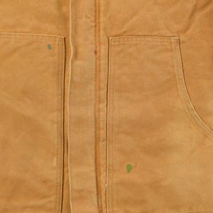 Puede incluir: Primer plano de una chaqueta de trabajo de color marr&oacute;n con dos bolsillos delanteros grandes y una cremallera central. La chaqueta tiene un aspecto desgastado con costuras visibles y peque&ntilde;as manchas de pintura. La tela parece ser un material duradero y resistente.