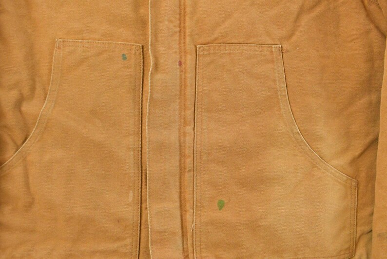 Puede incluir: Primer plano de una chaqueta de trabajo de color marr&oacute;n con dos bolsillos delanteros grandes y una cremallera central. La chaqueta tiene un aspecto desgastado con costuras visibles y peque&ntilde;as manchas de pintura. La tela parece ser un material duradero y resistente.