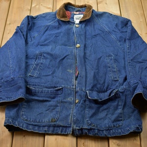 Puede incluir: Chaqueta vaquera azul con cuello de pana marrón y dos bolsillos delanteros grandes. La chaqueta tiene cierre de botones y forro acolchado. Las mangas tienen puño abotonado. La chaqueta está tendida sobre una superficie de madera.