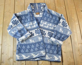 May include: A blue and white patterned cardigan sweater with a shawl collar. The sweater features a repeating pattern of deer, trees, and geese. The cuffs are rolled up. The sweater is made of a soft, warm material.