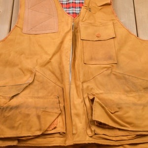 May include: A brown canvas hunting vest with multiple pockets and a zipper closure. The vest has a plaid lining and a leather patch on the back.