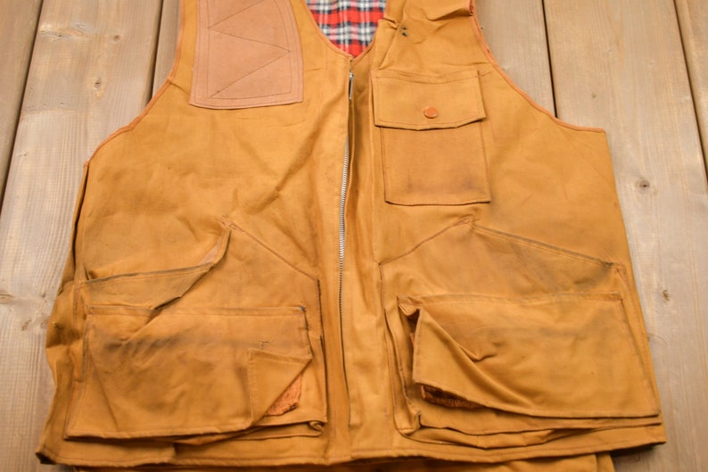 May include: A brown canvas hunting vest with multiple pockets and a zipper closure. The vest has a plaid lining and a leather patch on the back.
