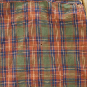 May include: Close-up of a plaid shirt in shades of orange, green, blue, and brown. The fabric is woven in a classic plaid pattern, with intersecting lines creating a grid-like design. The shirt's collar and part of the sleeve are visible.