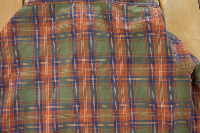 May include: Close-up of a plaid shirt in shades of orange, green, blue, and brown. The fabric is woven in a classic plaid pattern, with intersecting lines creating a grid-like design. The shirt's collar and part of the sleeve are visible.