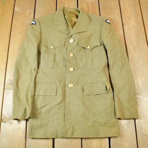 Puede incluir: Chaqueta militar verde oliva con cuatro bolsillos delanteros y botones dorados. La chaqueta tiene parches en los hombros y un cuello clásico. La chaqueta se exhibe sobre una superficie de madera.