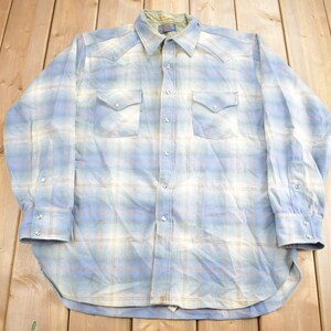 Puede incluir: Una camisa de manga larga de cuadros azules y blancos con cuello abotonado y dos bolsillos en el pecho.