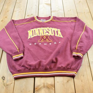 May include: A maroon long-sleeve sweatshirt featuring the text MINNESOTA in yellow letters above a gold and maroon M logo and the word GOPHERS in white. The garment has yellow and white striped ribbed cuffs, a striped hem, and yellow piping.