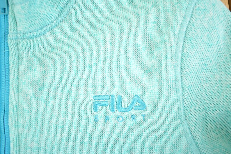 May include: Close-up of a light teal knit jacket with a full zipper. The jacket features the embroidered FILA SPORT logo in a matching teal color. The knit fabric has a speckled appearance.