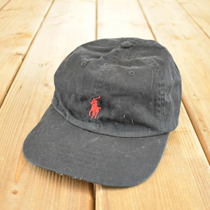 Op de afbeelding: Zwarte baseballcap met een gebogen klep. De cap heeft een rood geborduurd logo van een polospeler op de voorkant. De cap is gemaakt van een zachte, versleten stof en heeft een knoop aan de bovenkant.