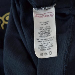 May include: A white care tag with black text that reads "Johnson Motors, Inc." and includes care instructions for the garment. The tag is attached to a dark blue t-shirt.
